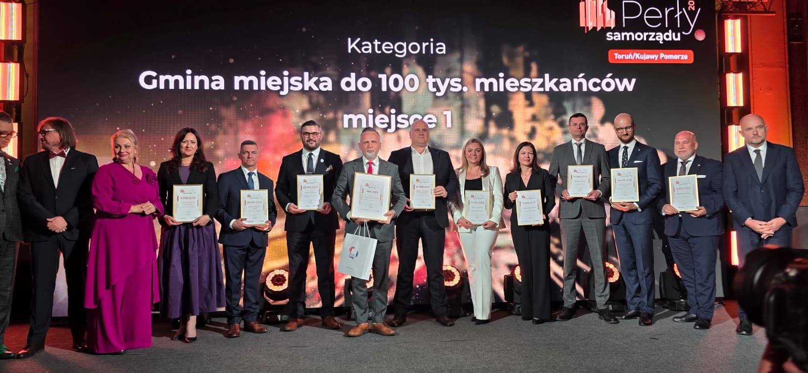 Czternaście osób stoi w rzędzie na scenie wewnątrz sali. Każda osoba ma eleganckie, formalne ubranie i trzyma w rękach ramki z dyplomami. W tle znajduje się duży podświetlany ekran i ciepłe, pomarańczowo-czerwone oświetlenie.