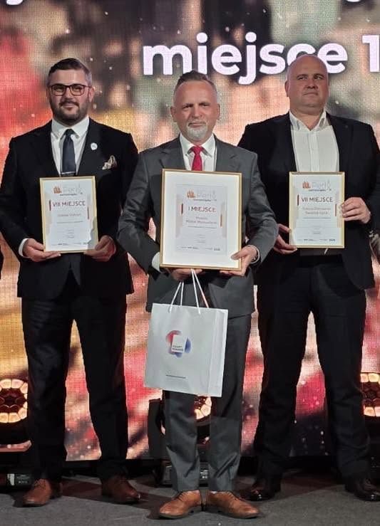 Trzech mężczyzn w garniturach stoi na scenie i trzyma oprawione dyplomy. Środkowy mężczyzna trzyma dodatkowo papierową torbę. Tło to podświetlany ekran z rozmytymi, ciepłymi kolorami.