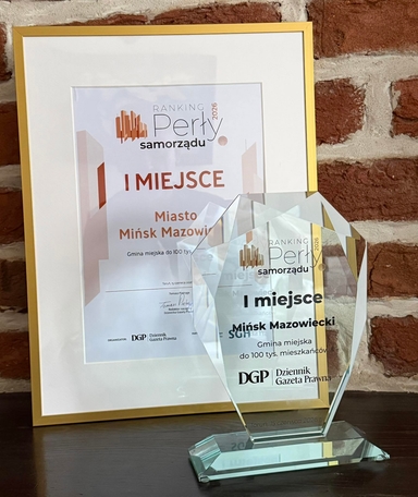 Złota ramka z dyplomem wisząca na czerwonej ceglanej ścianie. Przed ramką stoi przezroczysty kryształowy puchar na ciemnym drewnianym blacie.