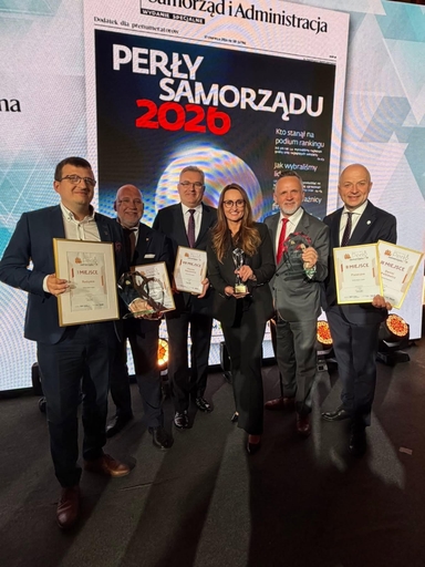 Sześć osób stoi na scenie wewnątrz przed dużym czarnym i białym plakatem z napisem „PERŁY SAMORZĄDU 2026”. Osoby mają formalne ubrania i krawaty. Trzymają ramki z dyplomami i szklane statuetki. Tło ma jasne geometryczne wzory i sceniczne oświetlenie.