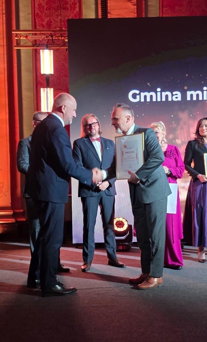Dwóch mężczyzn w garniturach ściska ręce na scenie podczas wręczania nagrody. Odbiorca trzyma ramkę z dyplomem. W tle stoją trzy osoby w eleganckich strojach i duży ekran z częściowo widocznym napisem, ciepłe czerwone oświetlenie, wnętrze sali.