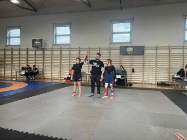 W hali sportowej na macie do walki stoi trójka osób: sędzia podnosi w górę rękę dziewczynki stojącej po lewej, druga dziewczynka stoi po prawej stronie. W tle widać ławki i osoby siedzące przy stolikach oraz drabinki gimnastyczne na ścianie.