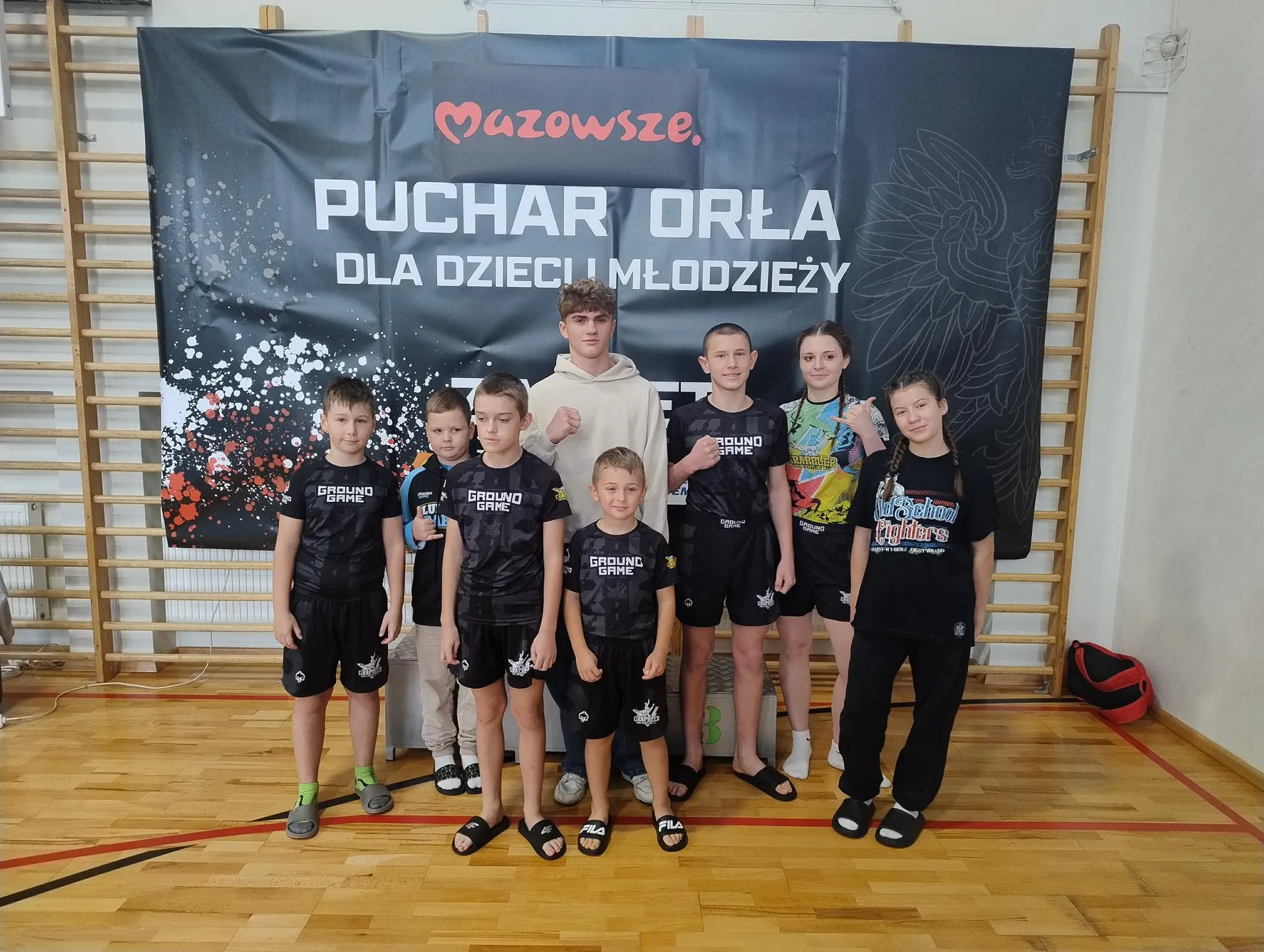 Grupa ośmiu dzieci i młodzieży stoi na drewnianej podłodze w hali sportowej na tle czarnego baneru z napisem „Mazowsze Puchar Orła dla dzieci i młodzieży”. Większość z nich ma na sobie sportowe ubrania i klapki. Za nimi metalowa ścianka do wspinaczki.