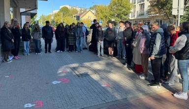 Grupa około trzydziestu osób stoi w skupieniu na chodniku przy ulicy w dzień.