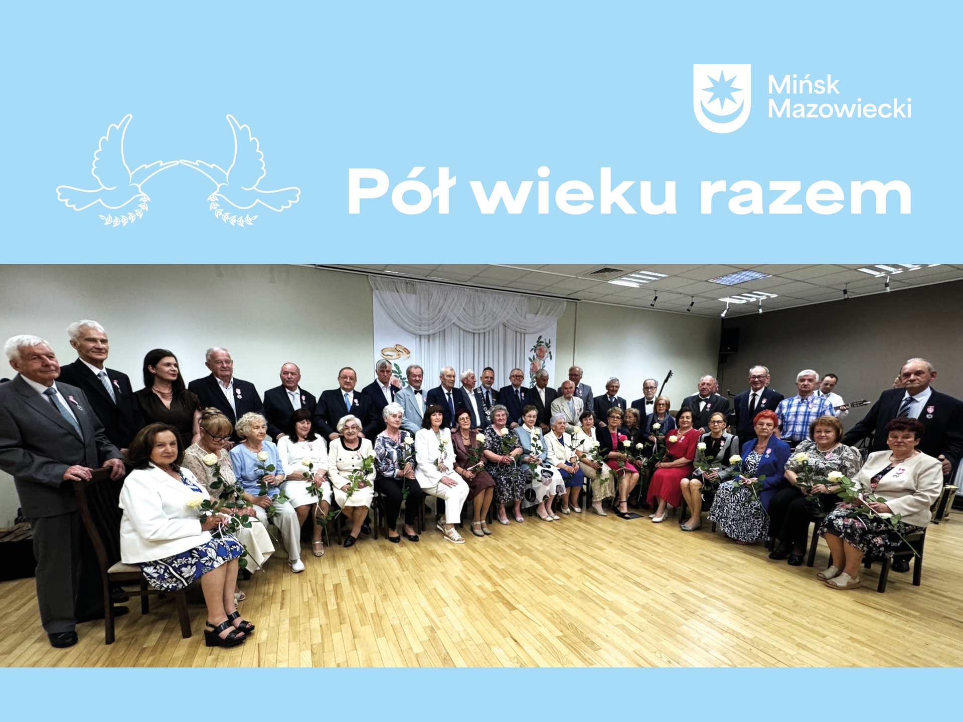 Duża grupa starszych osób siedzi i stoi w rzędach w sali, ustawiona do wspólnego zdjęcia. Wiele osób trzyma pojedyncze białe róże i ma przypięte medale. Na górze jasnoniebieski pas z białym napisem i herbem miasta oraz dekoracją gołębi. Podłoga drewniana, wnętrze oświetlone sufitowymi lampami.