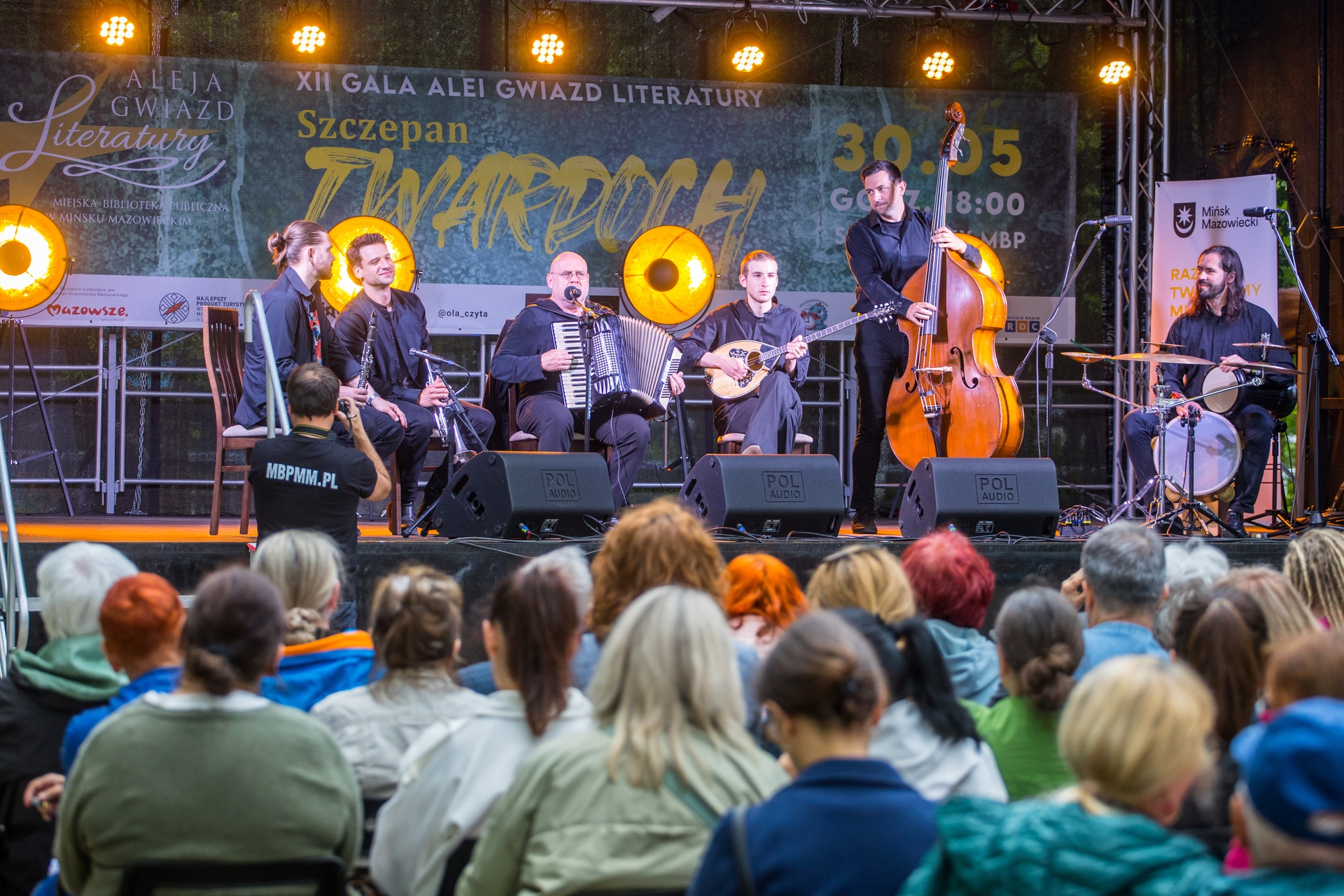 Sześciu muzyków gra na oświetlonej scenie na zewnątrz wieczorem. Publiczność siedzi na krzesłach na pierwszym planie. Na scenie akordeonista śpiewa, obok są klarnecista, gitarzysta lutniowy, kontrabasista stojący i perkusista. Tło tworzy duży baner i żółte reflektory.