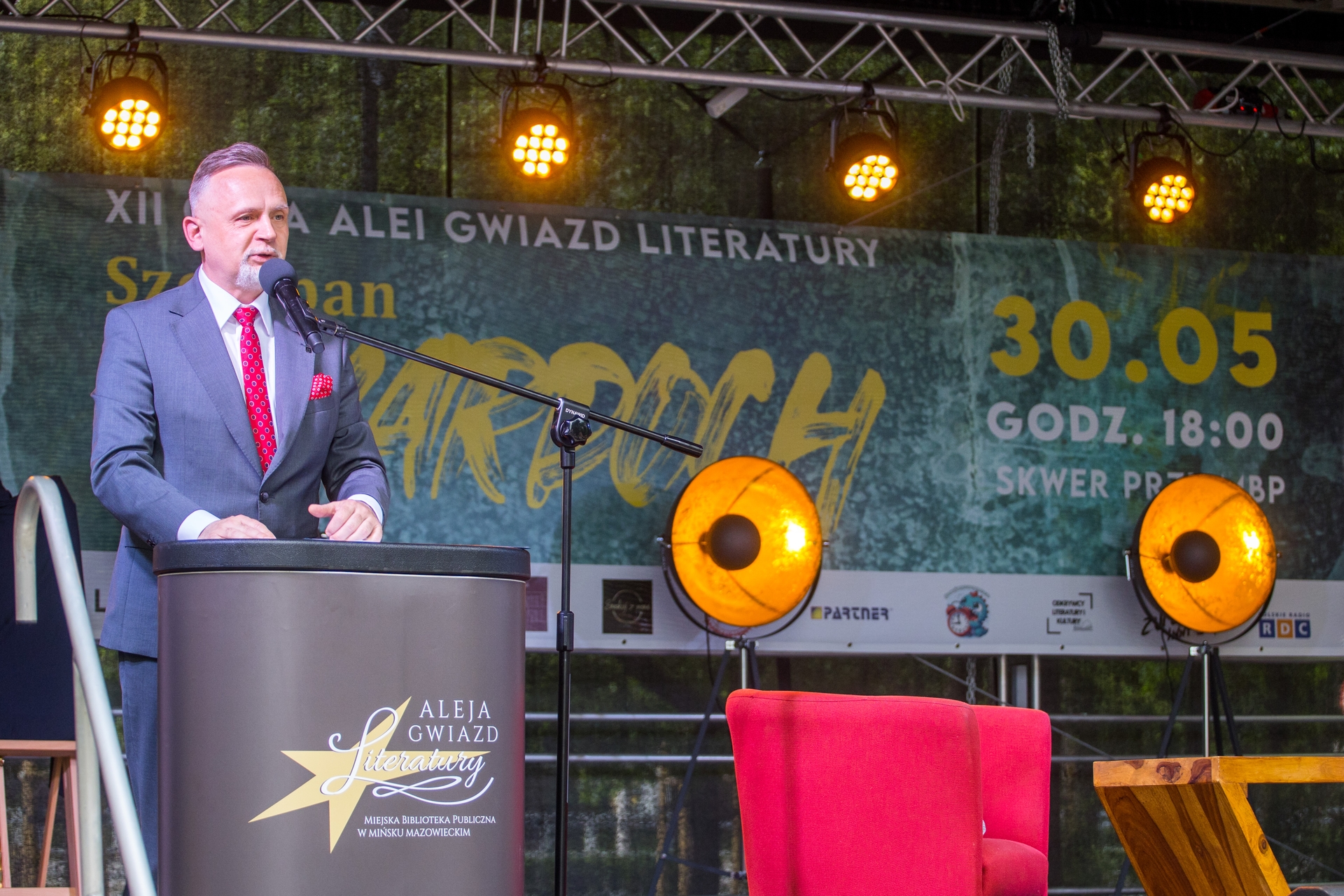 Mężczyzna w garniturze i czerwonym krawacie przemawia przy mównicy z mikrofonem na scenie. Za nim zielony baner i trzy żółte reflektory, obok stoi czerwony fotel.