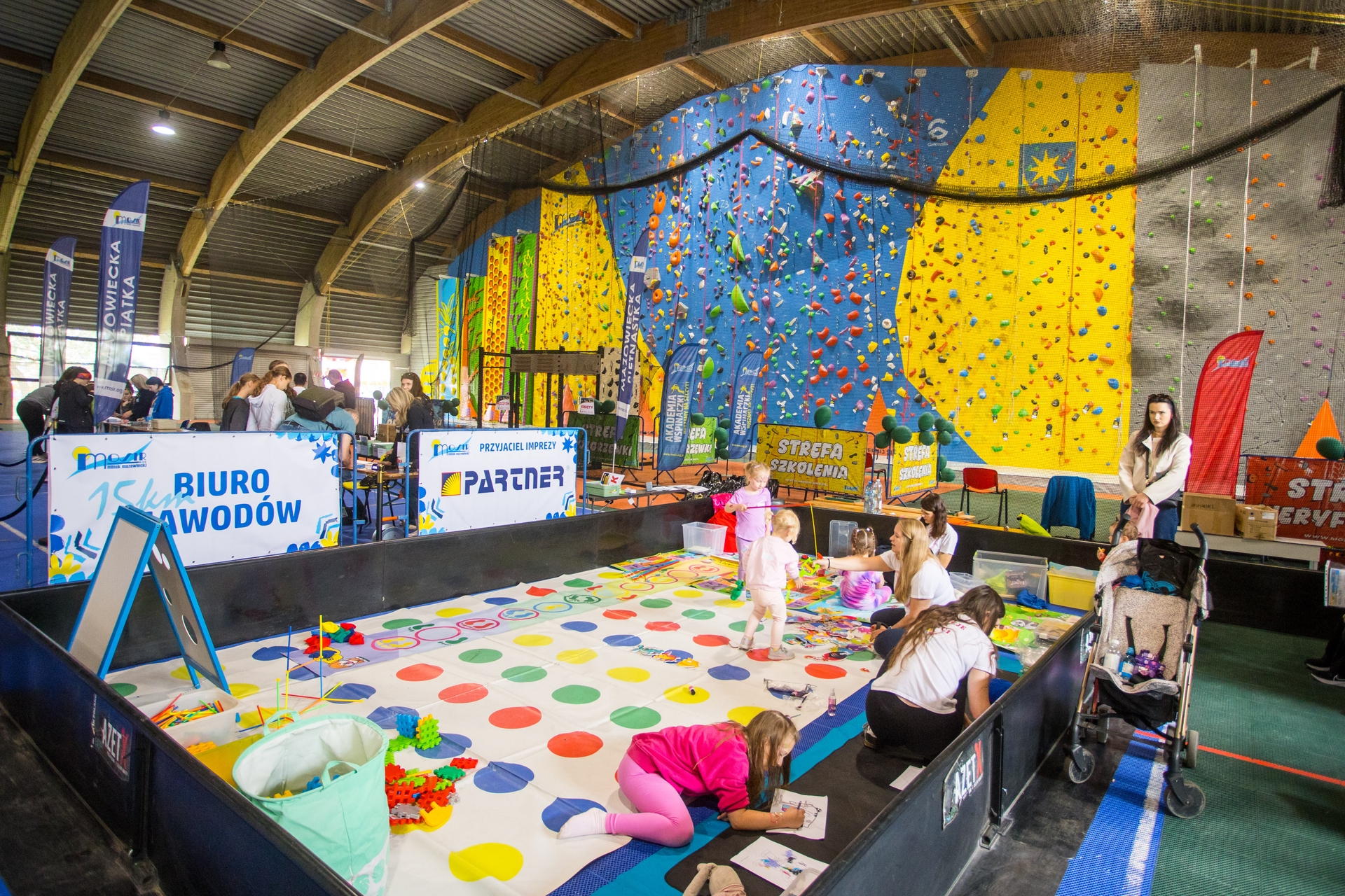Hala sportowa z wysoką, łukową konstrukcją dachu. Po prawej wielobarwna ścianka wspinaczkowa w niebiesko-żółtych panelach z kolorowymi chwytami. Na pierwszym planie strefa zabaw z dużą matą w kolorowe kółka, kilkoro dzieci i dorosłych bawiących się i rysujących, wózek dziecięcy po prawej.
