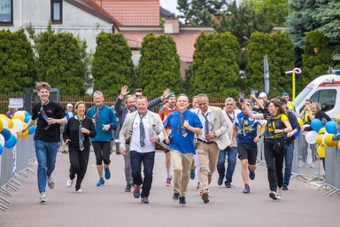 Grupa około piętnastu osób biegnie w stronę obserwatora po brukowanym deptaku na zewnątrz w ciągu dnia. Po bokach stoją metalowe bariery i kolorowe balony, w tle wysokie zielone żywotniki i zabudowania mieszkalne.