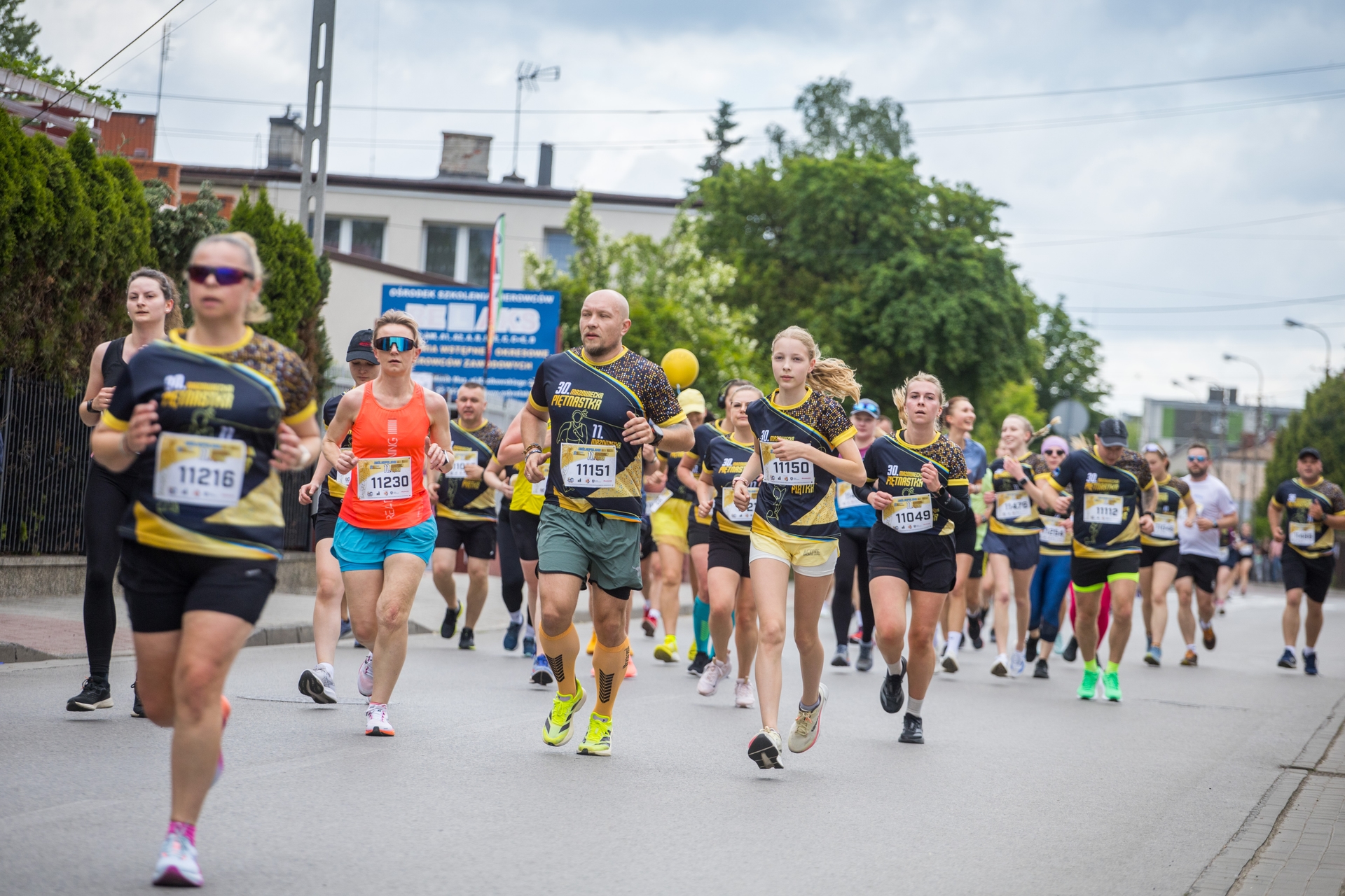 Grupa biegaczy biegnie ulicą w dzień. Większość ma czarne koszulki z żółtymi akcentami i numery startowe, w tle budynki, drzewa i pochmurne niebo.