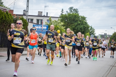 Grupa biegaczy biegnie ulicą w dzień. Większość ma czarne koszulki z żółtymi akcentami i numery startowe, w tle budynki, drzewa i pochmurne niebo.