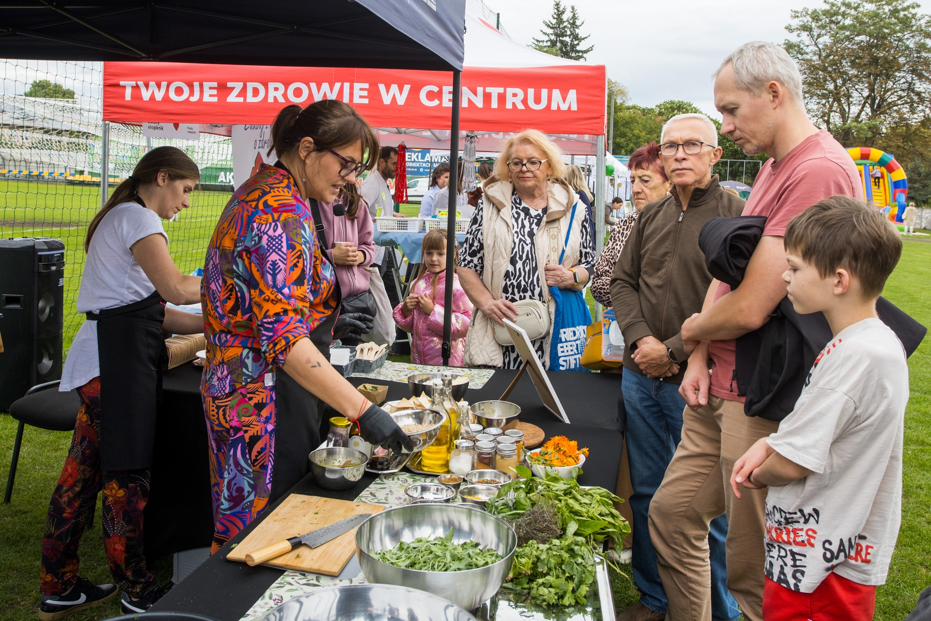 Stoisko gastronomiczne pod czerwonym pawilonem na zewnątrz na trawie. Kobieta w kolorowej sukience i rękawiczkach przygotowuje jedzenie przy stole z miskami, ziołami i butelkami oleju. Druga osoba w białym fartuchu pomaga obok. Przed stołem stoi grupa obserwatorów: dorośli i dwoje dzieci. W tle widoczna dmuchana zjeżdżalnia i drzewa.