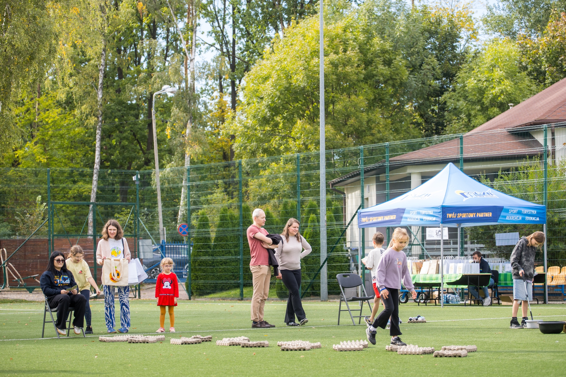 Ludzie na zewnętrznym boisku sportowym przy siatce i niskim ogrodzeniu. Kilka dzieci i dorosłych stoi i spaceruje obok niebieskiego namiotu, podłoże z zieloną trawą i ułożonymi kartonami po jajkach jako tor. W tle drzewa i budynek z dachem.