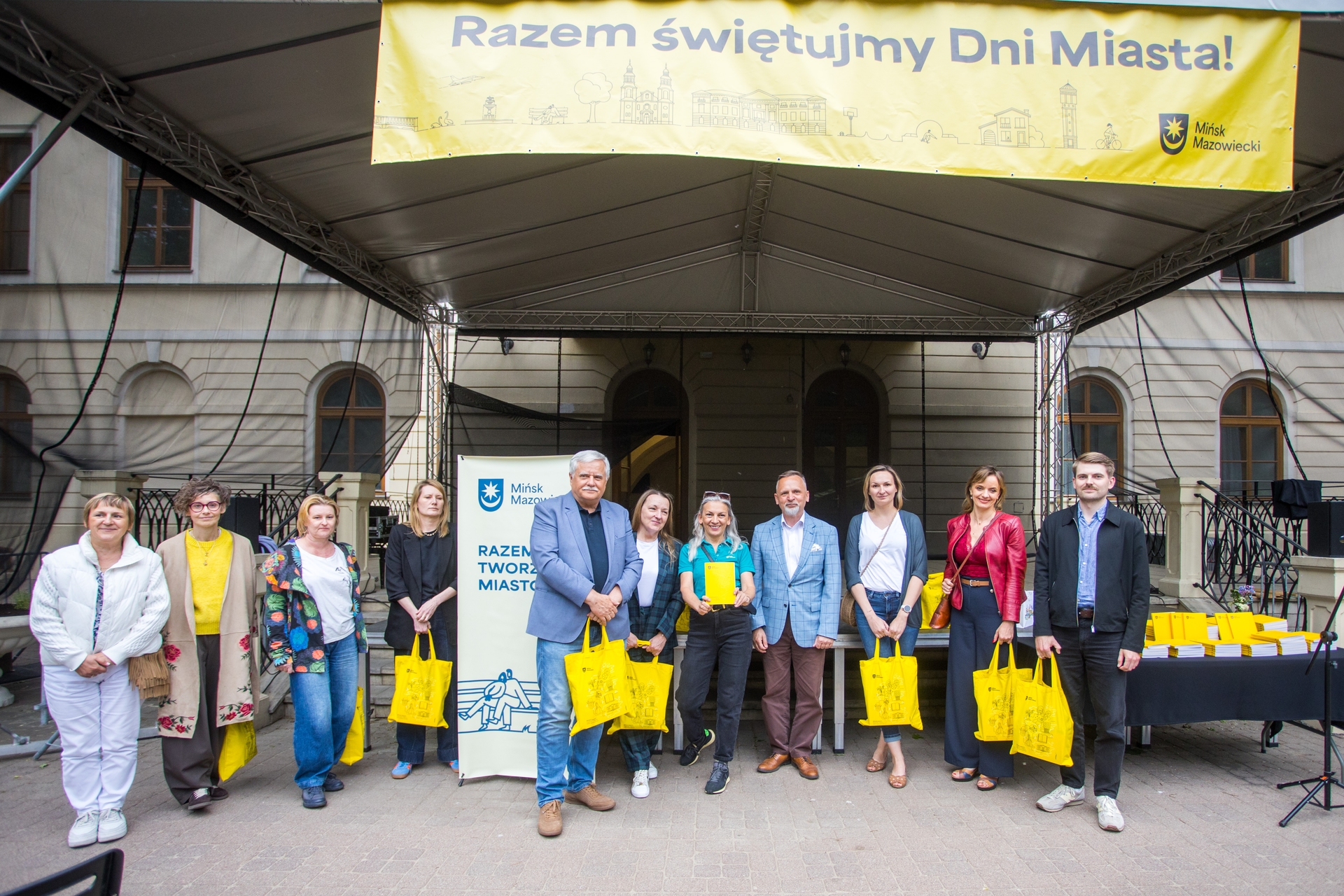 Grupa dwunastu osób stoi w rzędzie pod dużym namiotem na zewnętrznej scenie przed klasycznym budynkiem. Nad nimi wisi szeroki żółty baner, a większość osób trzyma żółte torby. Jest dzień, w tle widać arkady i okna fasady.