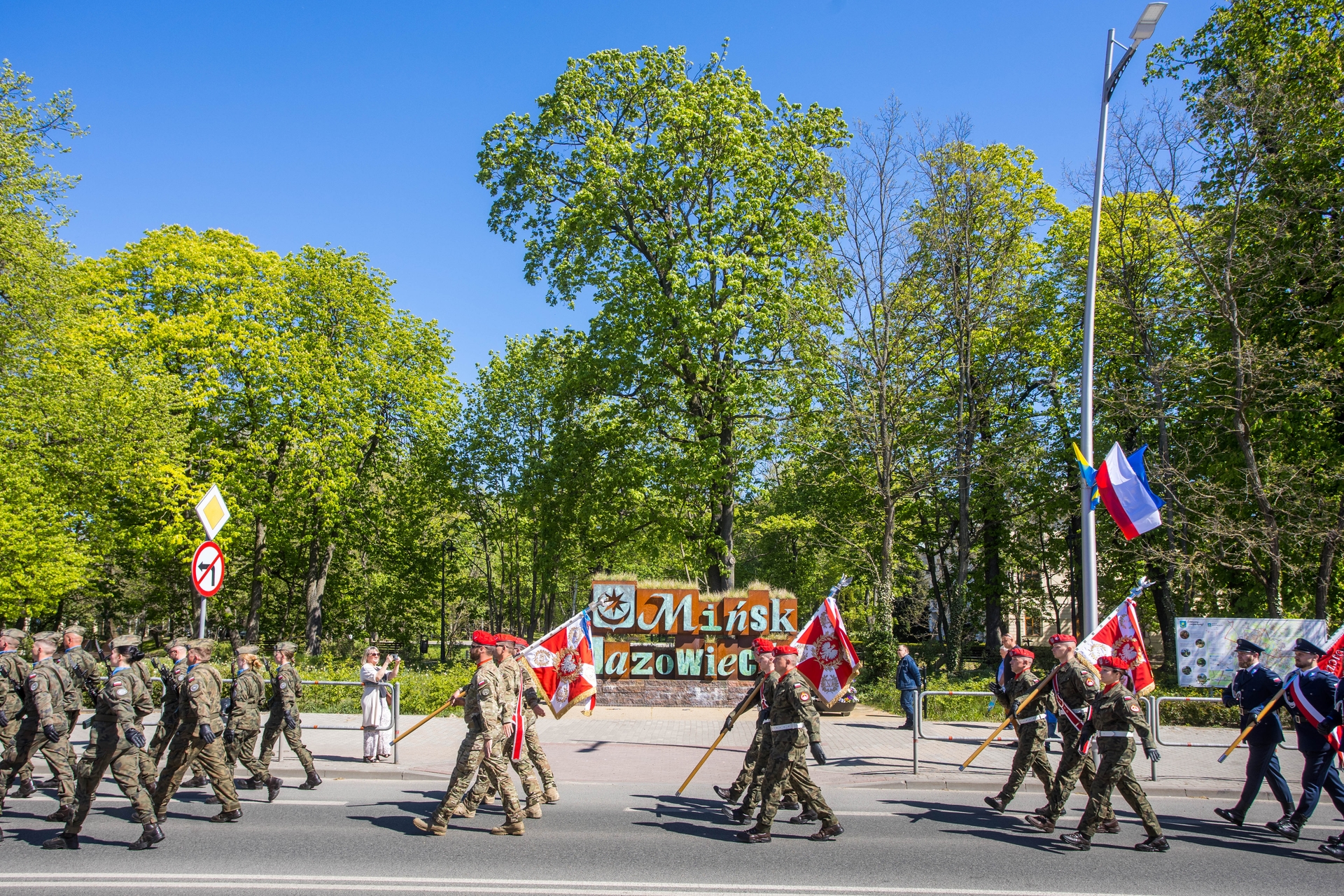 Żołnierze w mundurach maszerują w formacji wzdłuż ulicy niosąc czerwono-białe sztandary. Za nimi jest park z wysokimi zielonymi drzewami i jasnym błękitnym niebem. Przy chodniku stoi słup z zawieszonymi flagami i znaki drogowe.
