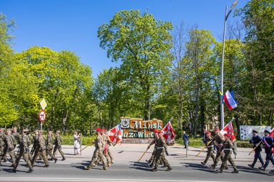 Żołnierze w mundurach maszerują w formacji wzdłuż ulicy niosąc czerwono-białe sztandary. Za nimi jest park z wysokimi zielonymi drzewami i jasnym błękitnym niebem. Przy chodniku stoi słup z zawieszonymi flagami i znaki drogowe.