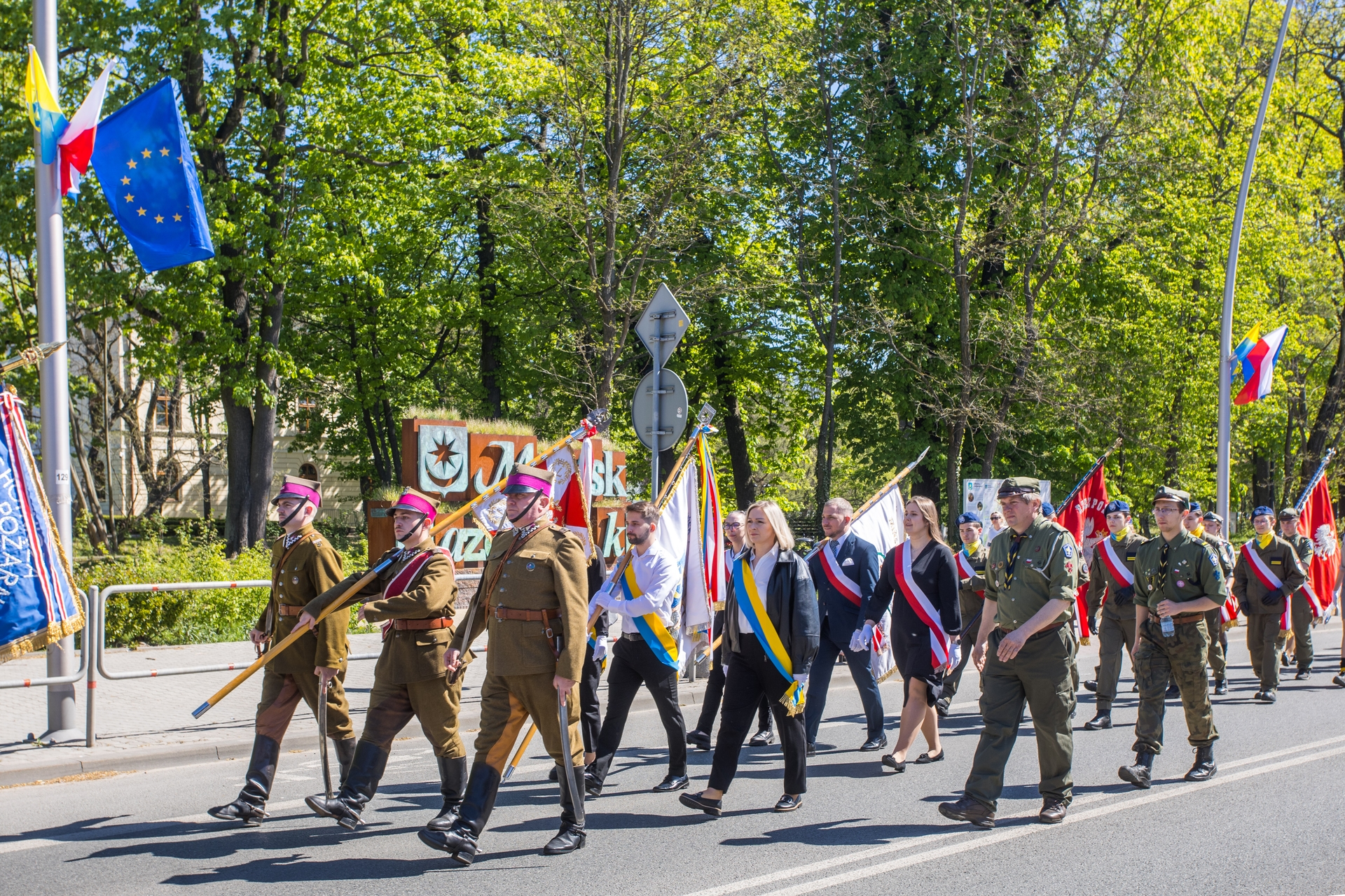 Grupa umundurowanych żołnierzy i cywilów maszeruje w uroczystej paradzie. Osoby niosą chorągwie i skrajne wstęgi, idą w świetle dnia wzdłuż ulicy; w tle zielone drzewa i widoczna flaga Unii Europejskiej.
