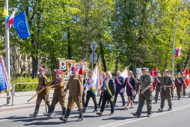 Grupa umundurowanych żołnierzy i cywilów maszeruje w uroczystej paradzie. Osoby niosą chorągwie i skrajne wstęgi, idą w świetle dnia wzdłuż ulicy; w tle zielone drzewa i widoczna flaga Unii Europejskiej.