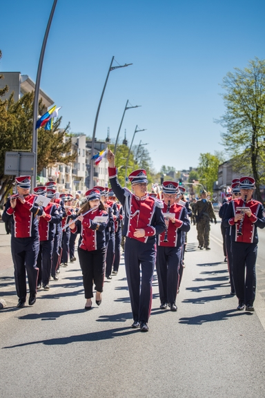 Orkiestra marszowa w czerwonych mundurach maszeruje środkową ulicą w słoneczny dzień; dyrygent na środku podnosi rękę. Muzycy grają na fletach i klarnetach, w tle niskie budynki, drzewa i flagi przy lampach ulicznych.