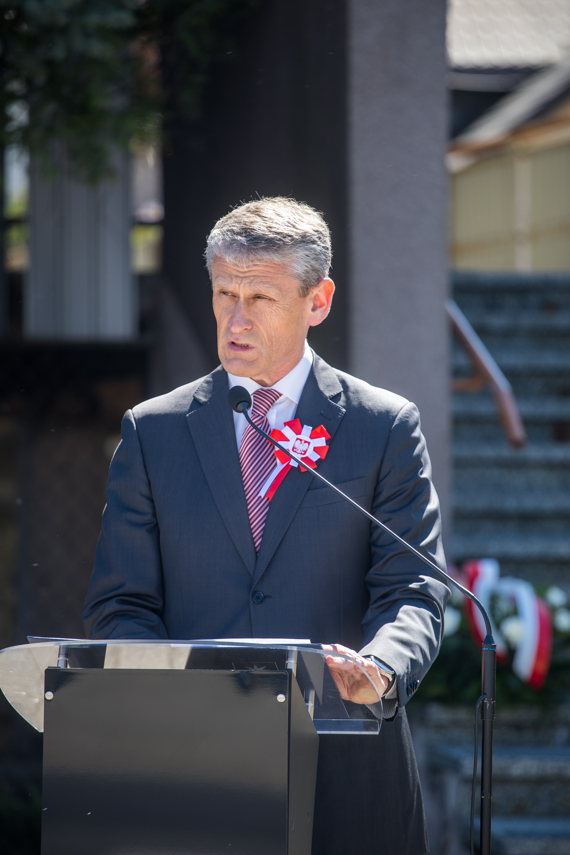 Mężczyzna w garniturze stoi przy pulpicie i przemawia do mikrofonu. Na marynarce ma biało-czerwoną kokardę. Scena odbywa się na zewnątrz w ciągu dnia, w tle schody i budynek.