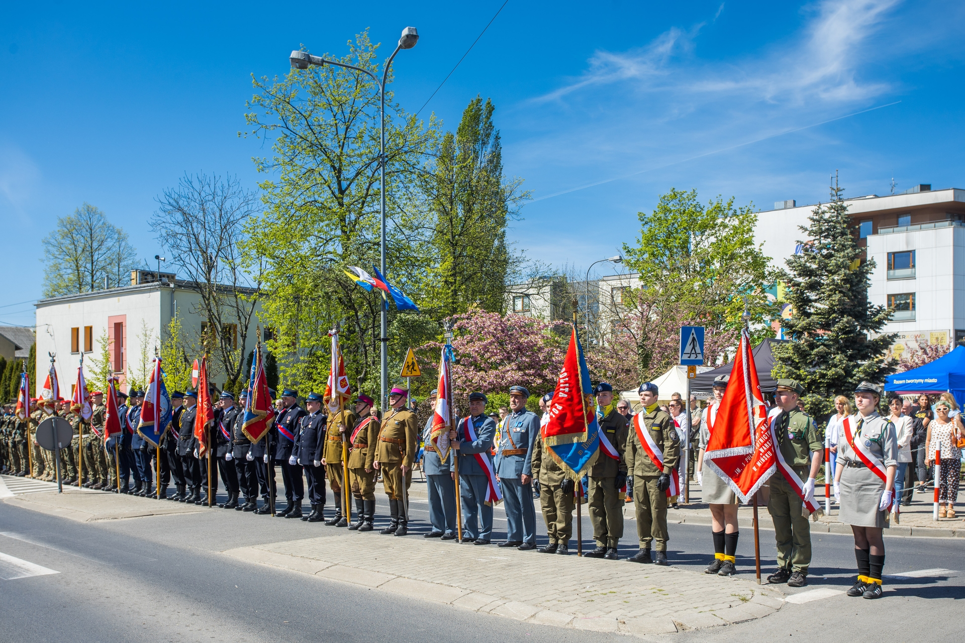 Grupa kilkudziesięciu umundurowanych osób stoi w szeregu na chodniku trzymając barwne sztandary. Dzień, jasne niebo, za nimi drzewa i budynki miejskie.