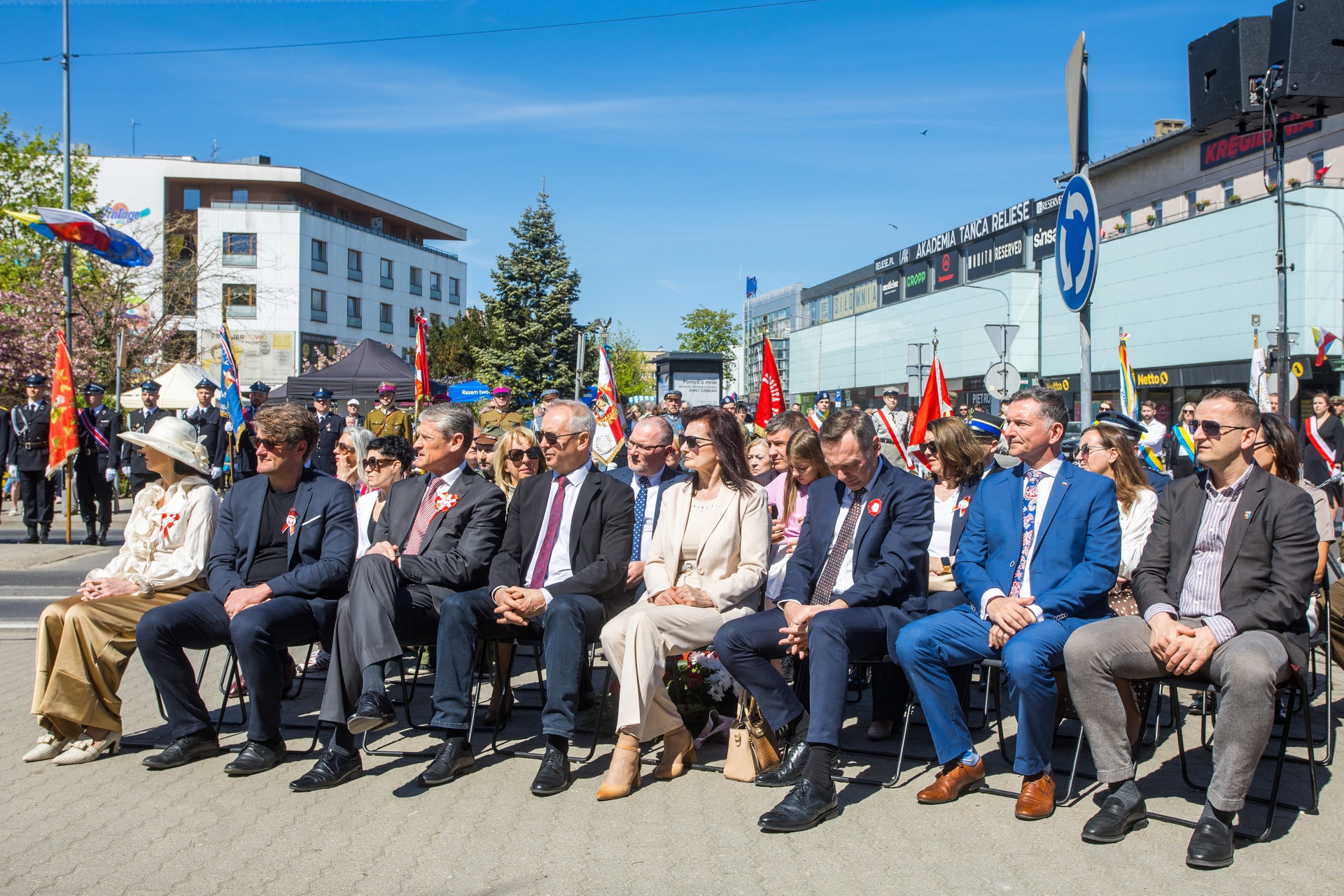Grupa kilkunastu osób siedzi w rzędzie na zewnątrz podczas uroczystości, większość w garniturach i formalnych strojach. Za nimi stoją umundurowani uczestnicy z chorągwiami i flagami. W tle niskie budynki, drzewa i jasne błękitne niebo.