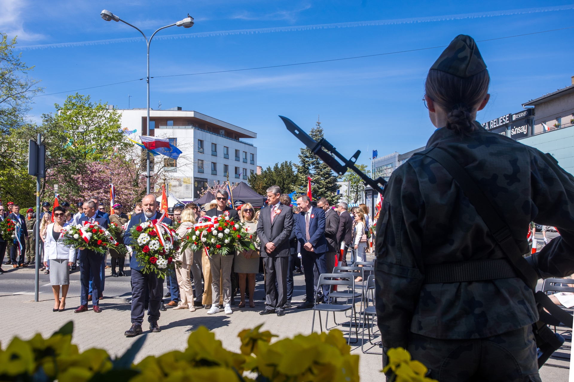 Dzień na zewnątrz podczas uroczystości przy miejskiej ulicy. Z przodu po prawej stoi żołnierz w mundurze widziany od tyłu z bagnetem przy ramieniu. Grupa osób w centrum niesie wieńce z czerwono-białymi wstęgami, za nimi budynki i powiewające flagi na tle niebieskiego nieba.