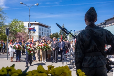 Dzień na zewnątrz podczas uroczystości przy miejskiej ulicy. Z przodu po prawej stoi żołnierz w mundurze widziany od tyłu z bagnetem przy ramieniu. Grupa osób w centrum niesie wieńce z czerwono-białymi wstęgami, za nimi budynki i powiewające flagi na tle niebieskiego nieba.