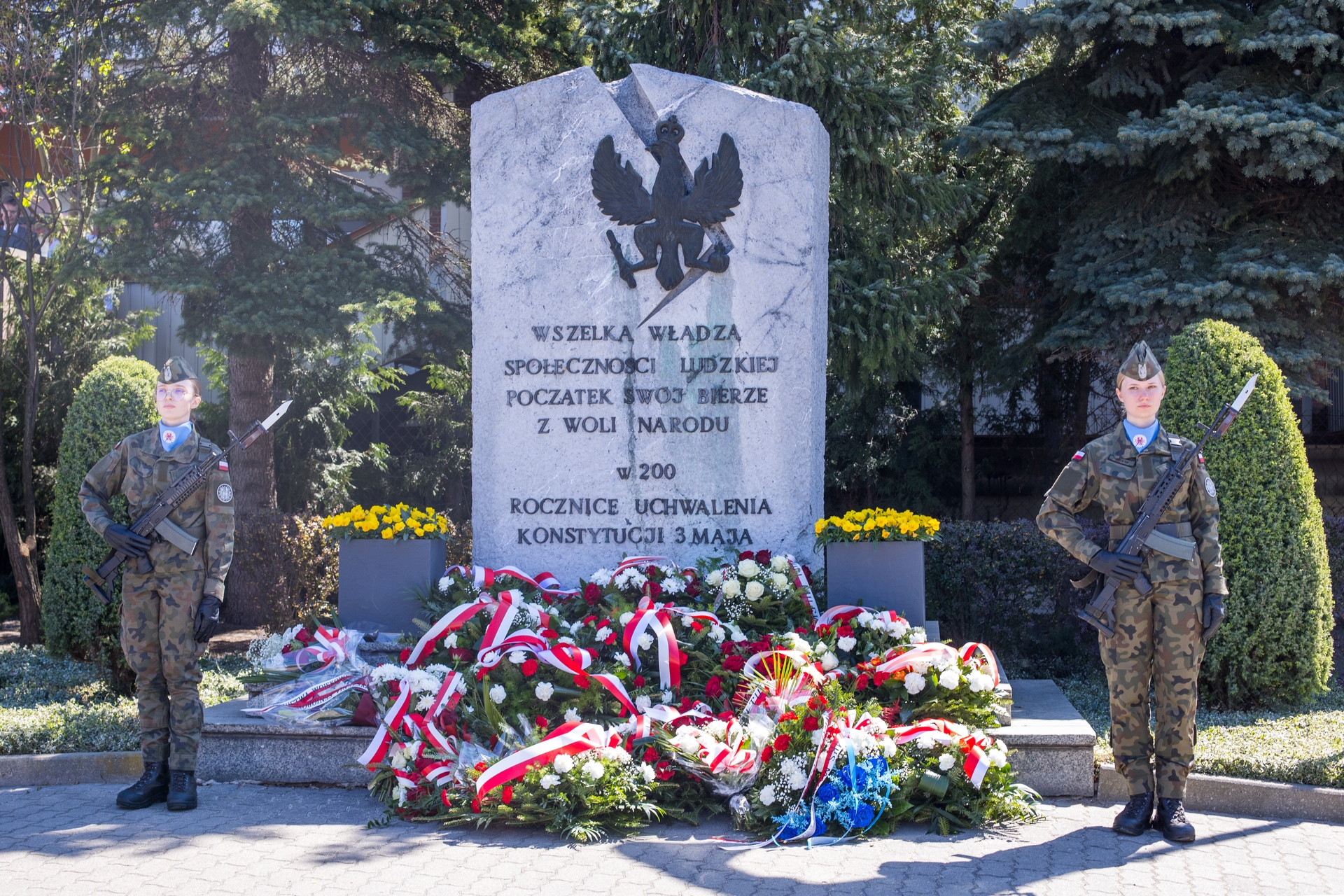 Kamienny pomnik z orłem państwowym stoi w centrum. Po obu stronach stoją dwie umundurowane żołnierki z karabinami w postawie honorowej. U podstawy leżą liczne wieńce i bukiety z biało-czerwonymi wstęgami. Scena odbywa się na zewnątrz w ciągu dnia, w tle wysokie iglaste drzewa.