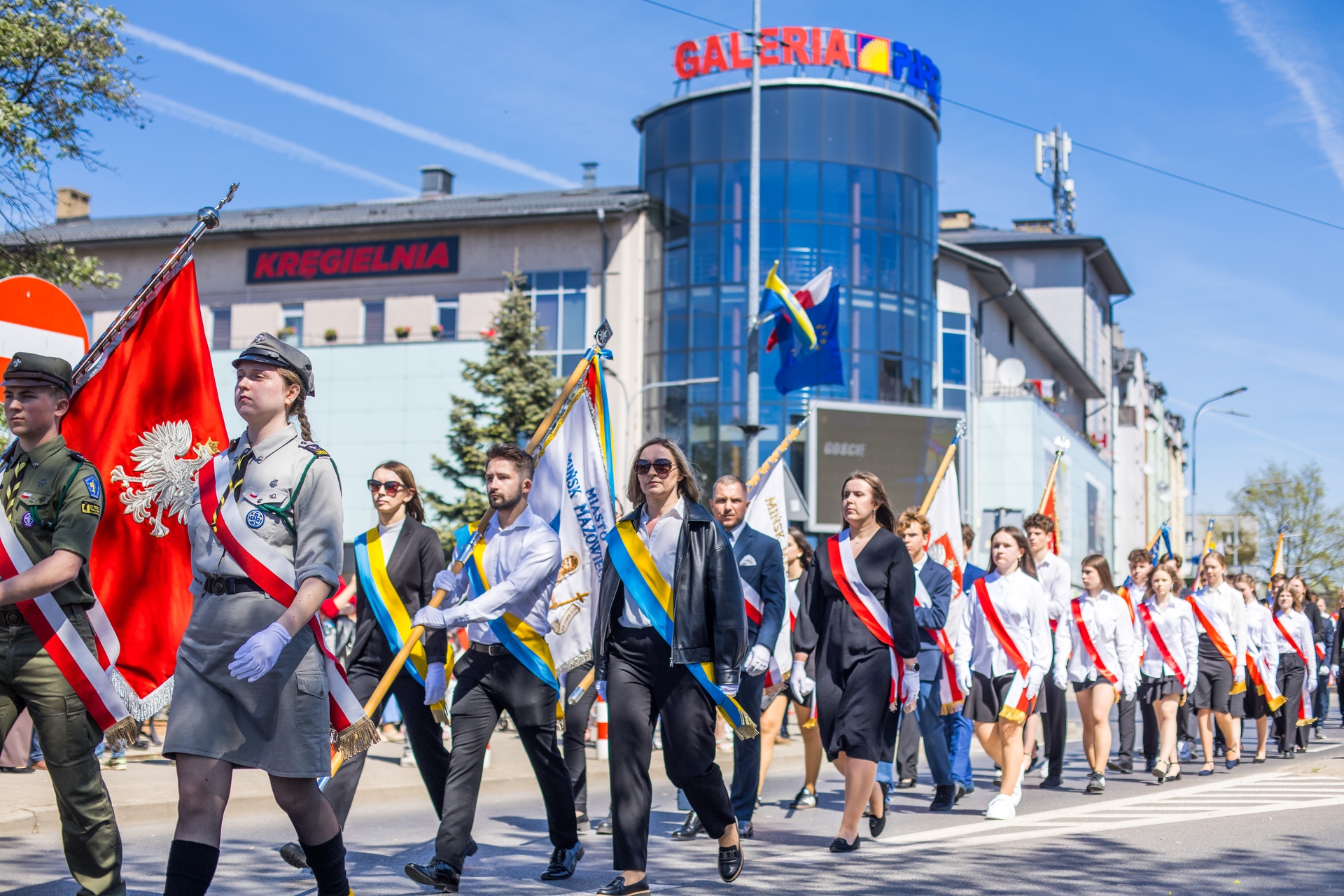 Grupa harcerzy, uczniów i dorosłych maszeruje w szyku po miejskiej ulicy. Osoby niosą kilkanaście kolorowych flag i proporców, niektórzy w mundurach, inni w strojach formalnych z szarfami. Jest dzień, niebo jest niebieskie, w tle widać nowoczesny, szklany budynek i kamienice.
