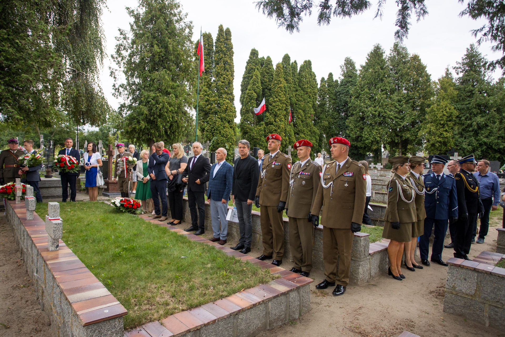 Grupa około trzydziestu osób stoi na cmentarzu na zewnątrz w ciągu dnia. Po prawej stoi kilku żołnierzy w mundurach z czerwonymi beretami, po lewej są cywile z wieńcami. W tle wysokie zielone drzewa i stojące polskie flagi.