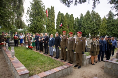 Grupa około trzydziestu osób stoi na cmentarzu na zewnątrz w ciągu dnia. Po prawej stoi kilku żołnierzy w mundurach z czerwonymi beretami, po lewej są cywile z wieńcami. W tle wysokie zielone drzewa i stojące polskie flagi.