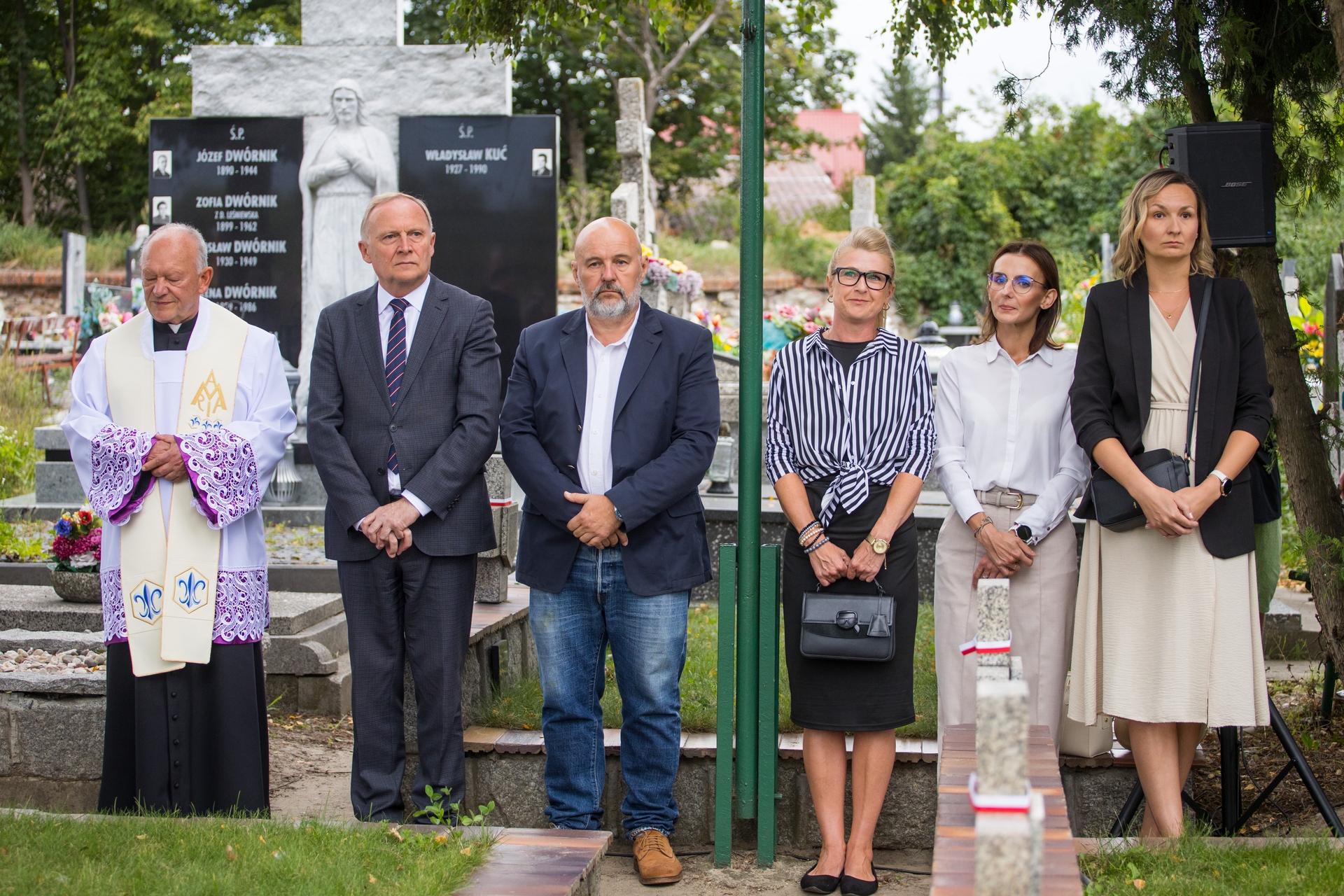 Sześć osób stoi na zewnątrz na cmentarzu przy nagrobkach i czarnym pomniku z rzeźbą, widać księdza po lewej i pięć osób w formalnych strojach ustawionych obok siebie, dzień, zielone drzewa w tle.