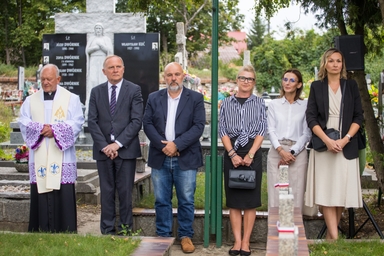 Sześć osób stoi na zewnątrz na cmentarzu przy nagrobkach i czarnym pomniku z rzeźbą, widać księdza po lewej i pięć osób w formalnych strojach ustawionych obok siebie, dzień, zielone drzewa w tle.