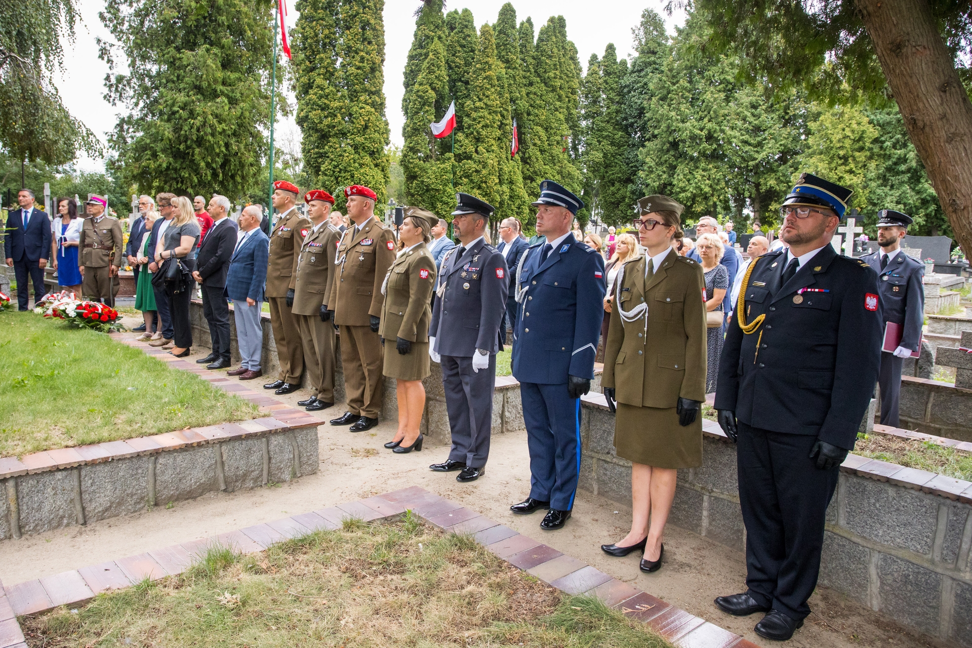 Kilkanaście osób w mundurach i ubraniach cywilnych stoi w równym szeregu na cmentarzu w ciągu dnia. W tle wysokie iglaste drzewa i kilka polskich flag na masztach. Na ziemi przy krawędzi alejki leżą wieńce z biało-czerwonymi kwiatami.