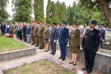Kilkanaście osób w mundurach i ubraniach cywilnych stoi w równym szeregu na cmentarzu w ciągu dnia. W tle wysokie iglaste drzewa i kilka polskich flag na masztach. Na ziemi przy krawędzi alejki leżą wieńce z biało-czerwonymi kwiatami.