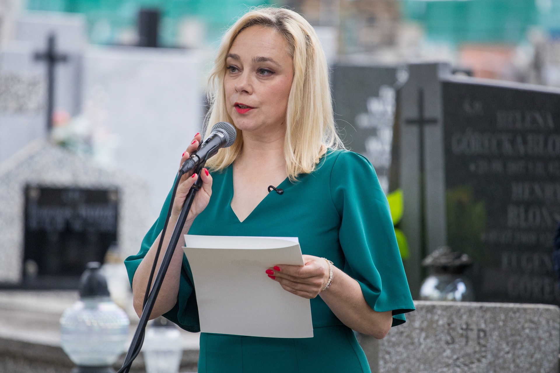 Kobieta o blond włosach mówi do mikrofonu trzymając kartki. Stoimy na zewnątrz przed nagrobkami i krzyżami, tło jest rozmyte.