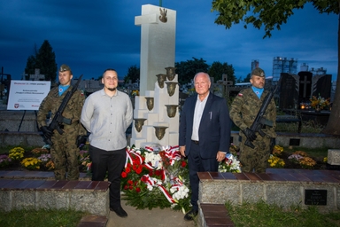 Dwie umundurowane osoby z bronią stoją na bokach pomnika w cmentarzu wieczorem. Dwaj cywile stoją przed centralnym kamiennym pomnikiem z metalowymi elementami. U podstawy leżą wieńce z biało-czerwonymi wstążkami, w tle groby i ciemnoniebieskie niebo.