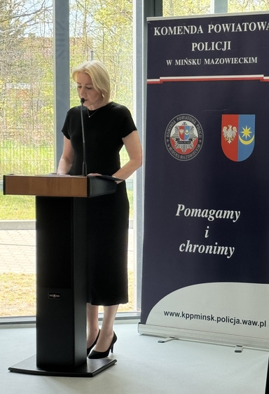 Kobieta stoi za mównicą i mówi do mikrofonu. Ubrana w czarną sukienkę i czarne szpilki. Wnętrze przy dużym oknie w dzień; obok stoi pionowy baner z herbami i emblematem policji.