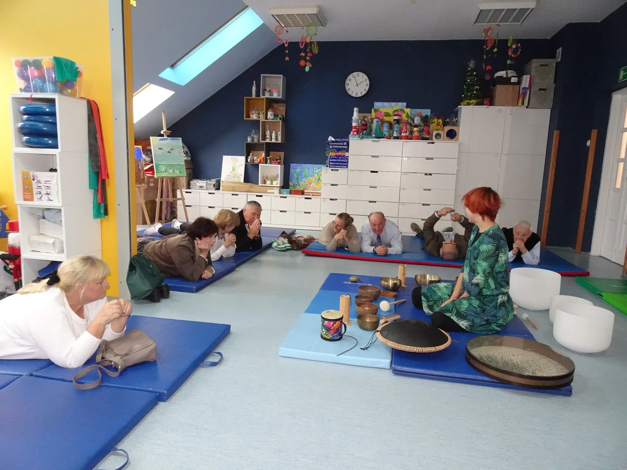 Grupa dorosłych leżących na matach w jasnym pokoju z ciemnoniebieską ścianą. Kobieta z krótkimi rudymi włosami siedzi na macie obok stołu z różnymi misami i instrumentami. W tle obrazy, półki z dekoracjami i zegar na ścianie.