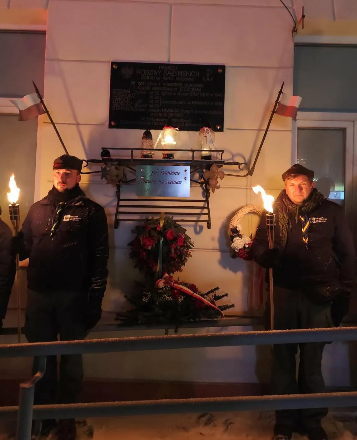 Dwóch mężczyzn stoi po obu stronach tablicy pamiątkowej na elewacji budynku. Każdy trzyma zapaloną pochodnię. Pod tablicą wiszą wieńce i zapalone znicze. Po bokach są małe flagi narodowe na drzewcach. Scena odbywa się na zewnątrz wieczorem przy sztucznym, ciepłym świetle; na ziemi widać śnieg.