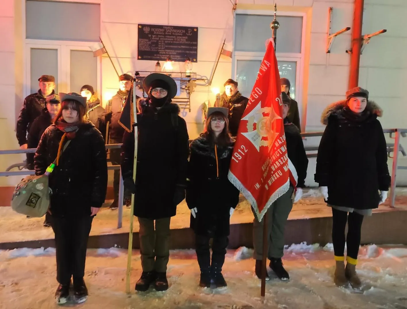 Pięć młodych osób stoi w rzędzie na zaśnieżonym chodniku przed oświetloną ścianą budynku. Środkowa osoba i osoba z prawej trzymają czerwony sztandar z emblematem i datą. W tle kilka osób stoi przy balustradzie pod tablicą pamiątkową. Scena dzieje się wieczorem, osoby noszą zimowe kurtki i czapki.