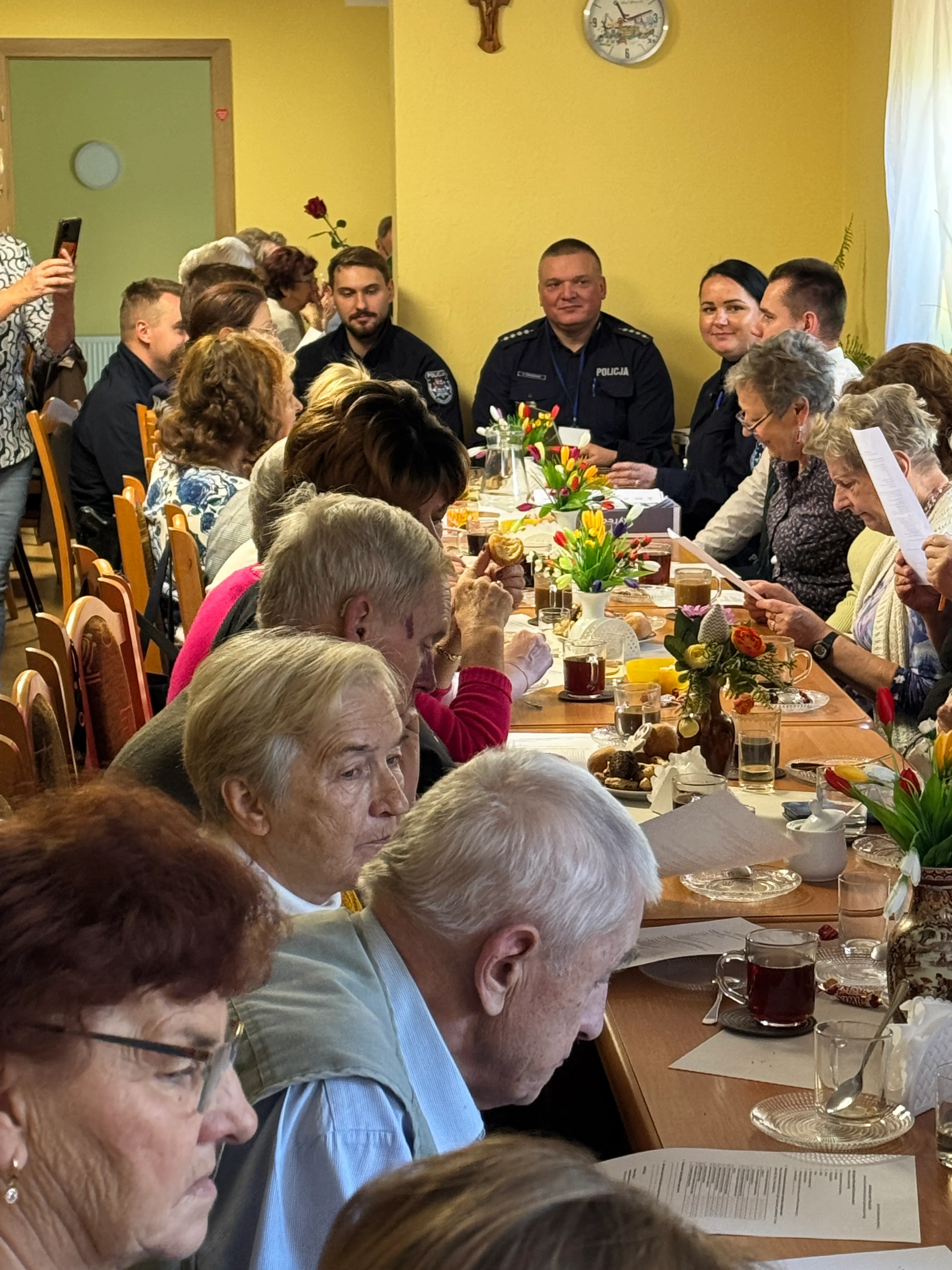 Grupa starszych osób siedzi przy długim stole w jasnym pomieszczeniu. Na stole leżą dokumenty, są napoje i dekoracje kwiatowe. W tle siedzą dwie osoby w mundurach policyjnych. Po lewej kobieta robi zdjęcie telefonem. Na ścianie żółtego koloru widoczny jest zegar i krucyfiks.