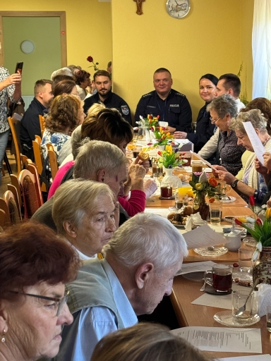 Grupa starszych osób siedzi przy długim stole w jasnym pomieszczeniu. Na stole leżą dokumenty, są napoje i dekoracje kwiatowe. W tle siedzą dwie osoby w mundurach policyjnych. Po lewej kobieta robi zdjęcie telefonem. Na ścianie żółtego koloru widoczny jest zegar i krucyfiks.