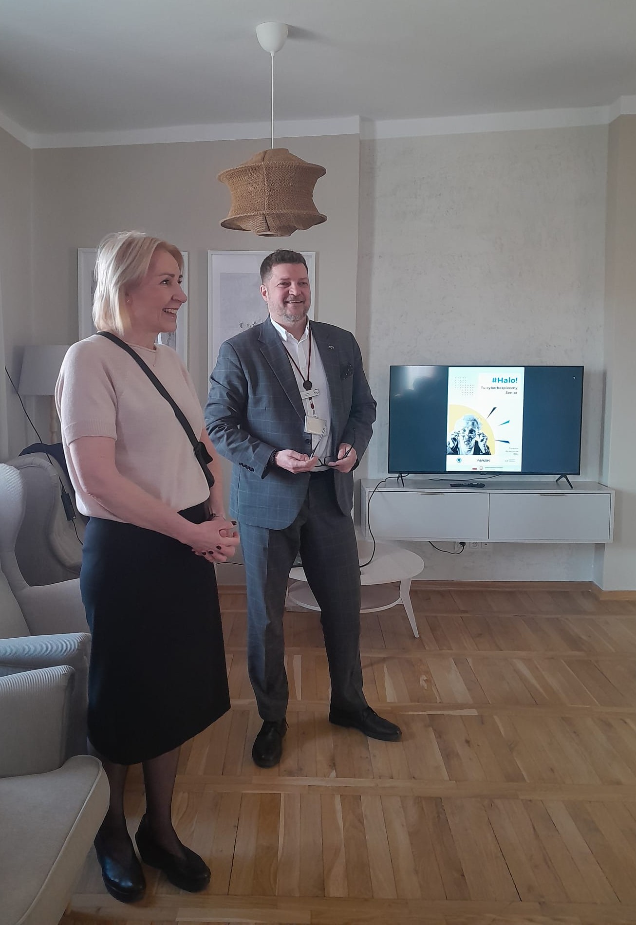 Dwoje dorosłych stoi w jasnym salonie. Kobieta po lewej ma jasne włosy i jasny sweter, mężczyzna po prawej ma garnitur i trzyma okulary. W tle niski biały stolik z telewizorem, beżowe ściany, drewniana podłoga i wisząca pleciona lampa.