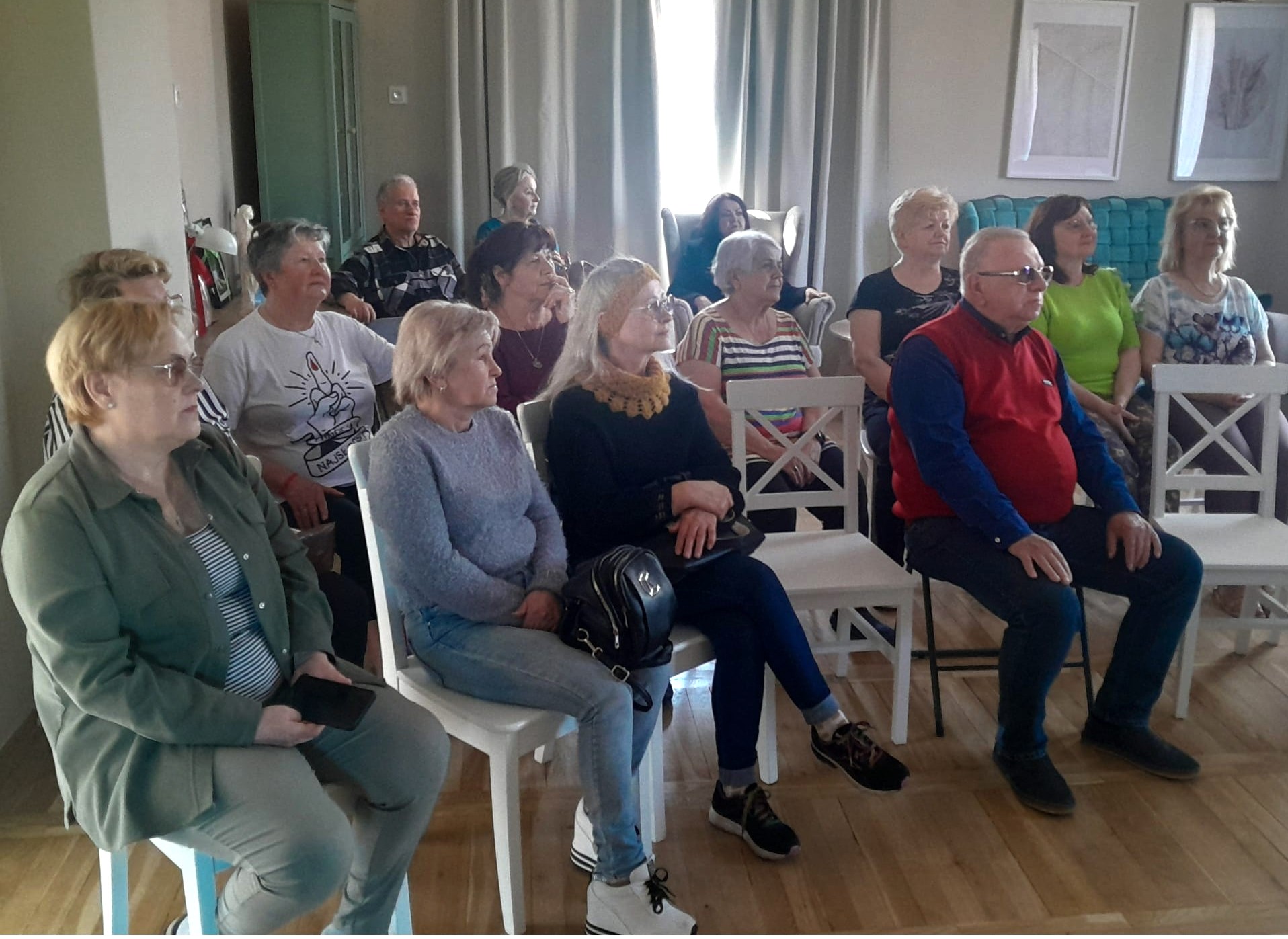 Kilkunastu dorosłych siedzi w rzędach białych krzeseł w jasnym, wewnętrznym pomieszczeniu z zasłonami i obrazami na ścianach; dominują starsze kobiety i kilku mężczyzn zwróconych ku przodowi.