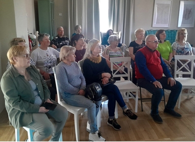 Kilkunastu dorosłych siedzi w rzędach białych krzeseł w jasnym, wewnętrznym pomieszczeniu z zasłonami i obrazami na ścianach; dominują starsze kobiety i kilku mężczyzn zwróconych ku przodowi.