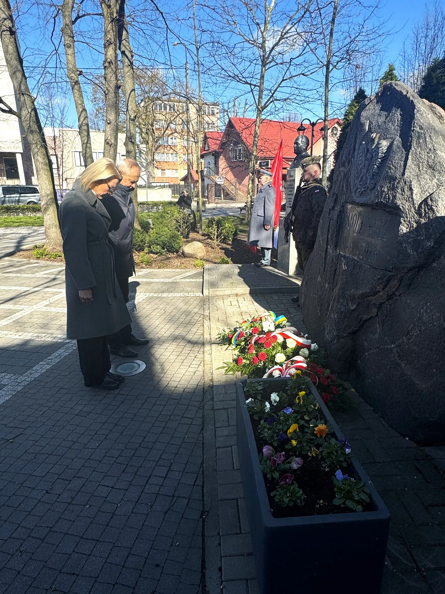 Dwie osoby stoją po lewej i pochylają głowy nad wieńcami złożonymi przed dużym kamiennym pomnikiem po prawej. Obok pomnika stoi żołnierz i mężczyzna w szarym płaszczu z czerwoną flagą. Na zewnątrz, dzień, w tle nagie drzewa i budynki.