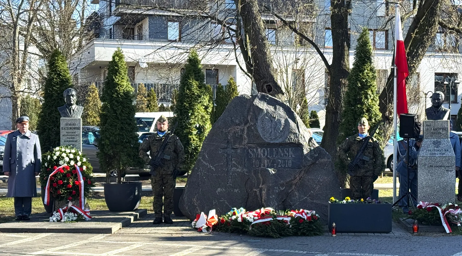 Kamienny pomnik z wyrytą inskrypcją Smoleńsk 10 IV 2010 stoi na środku placu. Przed nim leżą wiązanki i wieńce z biało-czerwonymi wstęgami. Po obu stronach stoją dwaj żołnierze w mundurach z bronią. Po bokach znajdują się marmurowe popiersia na postumentach. Po prawej widać maszt z biało-czerwoną flagą. Scena ma miejsce na zewnątrz w ciągu dnia, w tle drzewa i budynki.