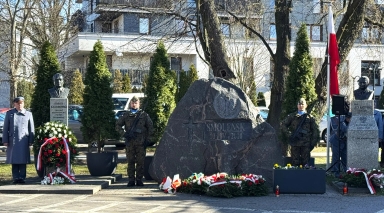 Kamienny pomnik z wyrytą inskrypcją Smoleńsk 10 IV 2010 stoi na środku placu. Przed nim leżą wiązanki i wieńce z biało-czerwonymi wstęgami. Po obu stronach stoją dwaj żołnierze w mundurach z bronią. Po bokach znajdują się marmurowe popiersia na postumentach. Po prawej widać maszt z biało-czerwoną flagą. Scena ma miejsce na zewnątrz w ciągu dnia, w tle drzewa i budynki.